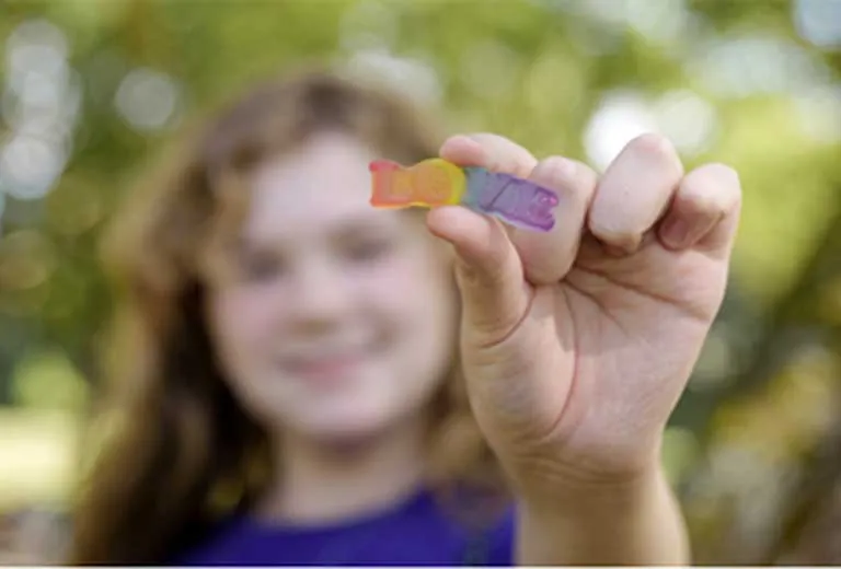 Eine junge Person hält ein regenbogenfarbenes Gummibärchen in die Kamera